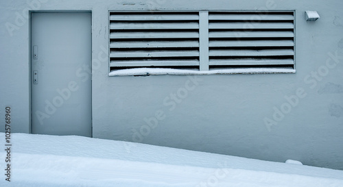 A building with a gray door, a slatted window, and a gray wall is covered in fresh snow.