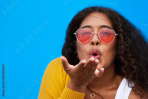 Woman, portrait and blow kiss in studio with heart sunglasses for valentine day, flirt gesture and romance. Girl, eyewear and love emoji, date inspiration and gratitude connection on blue background photo