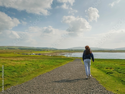 Teenager girl walking on a small country road. Stunning nature landscape in the background with green fields, blue ocean, mountains and cloudy sky. Downpatrick head, Ireland. Travel and tourism.