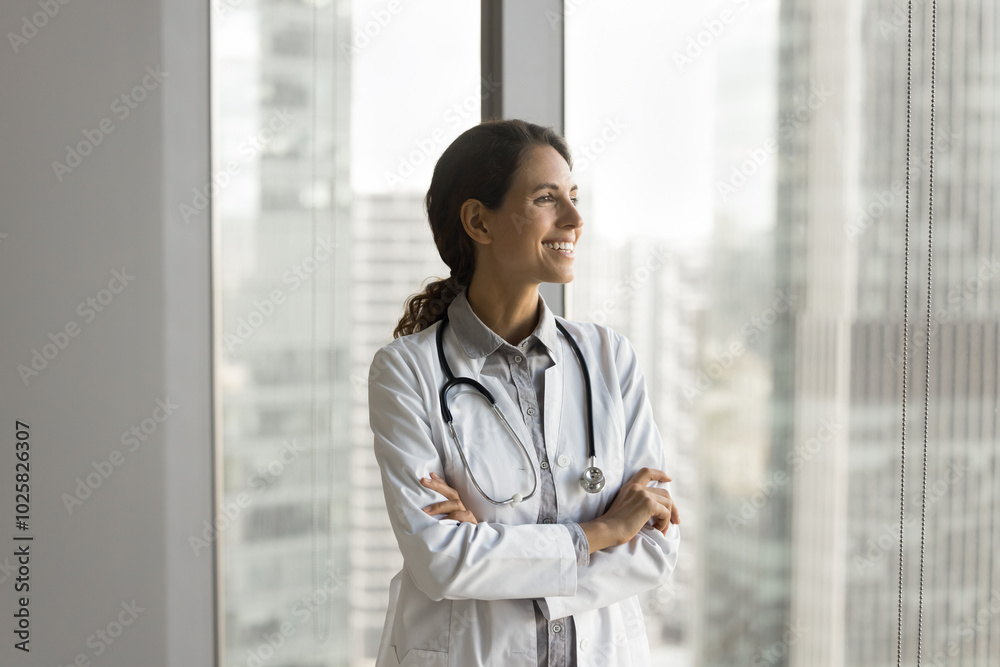 Happy young lady physician in charge stand by window at modern clinic office look at distance think about complete recovery of severe patient. Millennial female doc enjoy work visualize career success Happy young lady physician in charge stand by window at modern clinic office look at distance think about complete recovery of severe patient. Millennial female doc enjoy work visualize career success