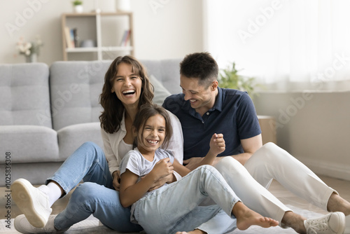 Sweet home. Laughing candid millennial spouses with preteen child mother father little daughter enjoy funny indoor activity messing tickling on floor with heating play in crazy naughty mood on weekend photo