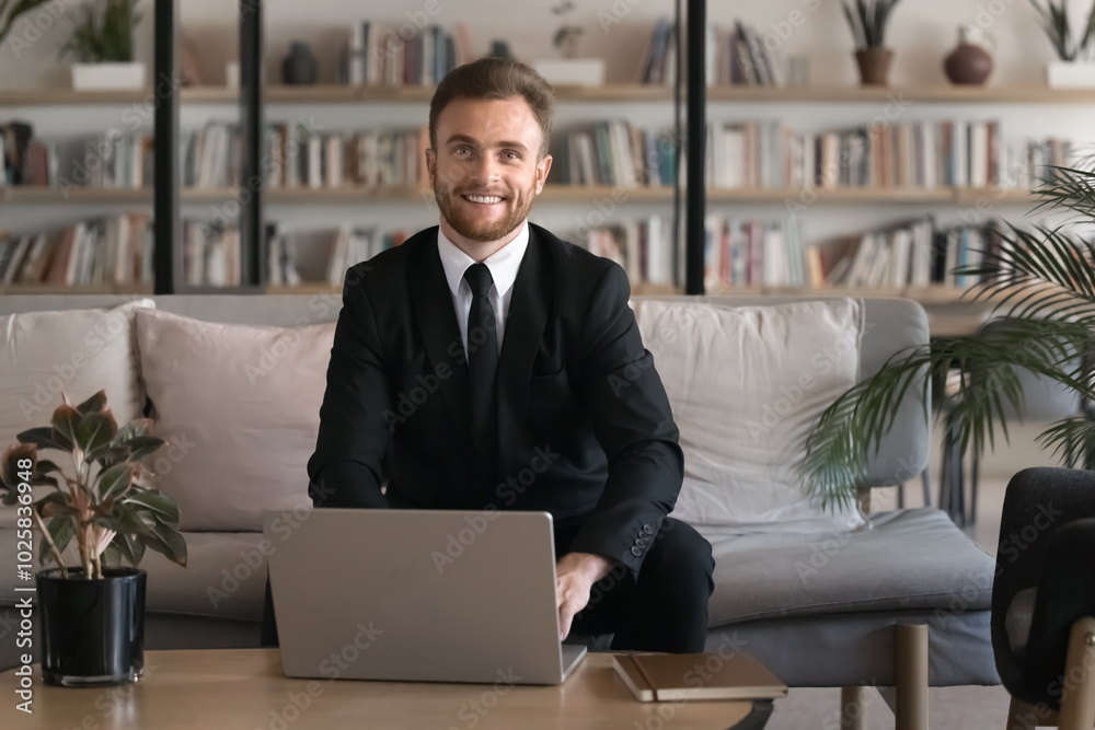 Happy successful young businessman in formal suit using laptop in modern co-working with library, sitting on couch, working on educational project, looking at camera, smiling for professional portrait Happy successful young businessman in formal suit using laptop in modern co-working with library, sitting on couch, working on educational project, looking at camera, smiling for professional portrait