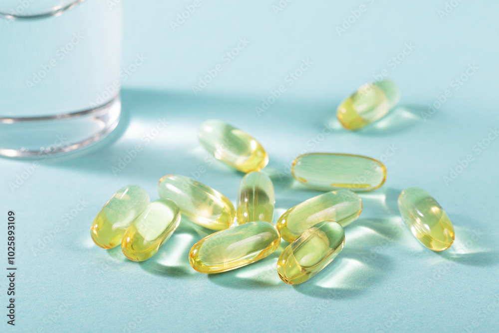 Diet, nutrition, healthy eating concept. Oil filled capsules of food supplements and glass of water for pouring yellow softgels capsules on light blue background.