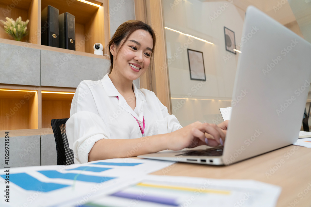 A woman is sitting at a desk with a laptop open in front of her. She is smiling and she is enjoying her work. The desk is cluttered with papers and books, and there is a potted plant in the background