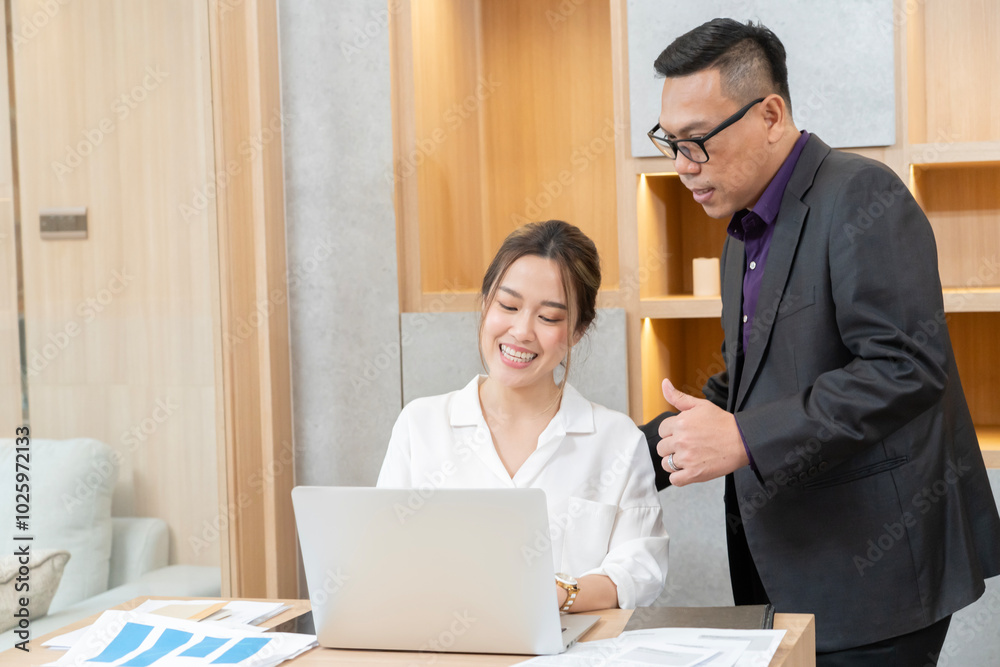 A man and a woman are sitting at a desk with a laptop. The man is pointing at the laptop screen
