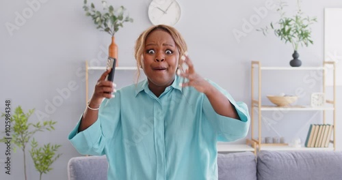 Happy excited african american woman in casual clothes holding mobile phone making yes winner's gesture celebrating success or win a lottery standing at home with money rain. 4k video.
