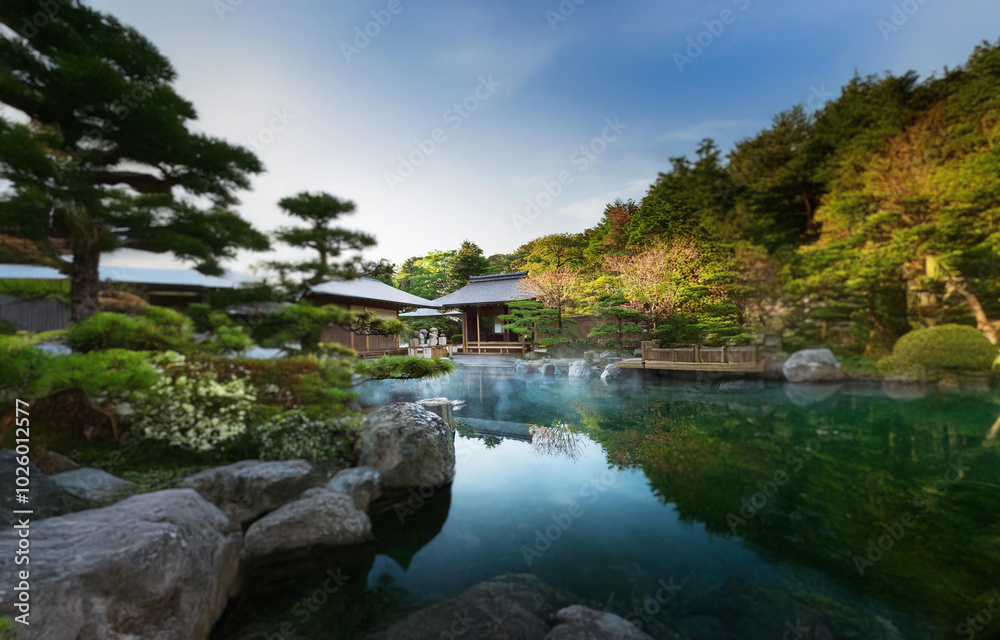 A tranquil Japanese garden features a serene pond with steam rising from its surface, surrounded by lush greenery and traditional wooden structures. A tranquil Japanese garden features a serene pond with steam rising from its surface, surrounded by lush greenery and traditional wooden structures.