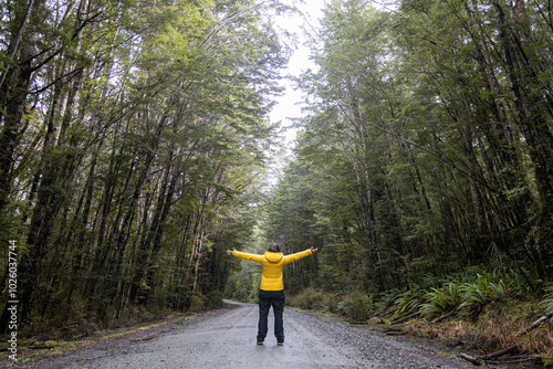 woman in yellow coat with arms open on path between trees