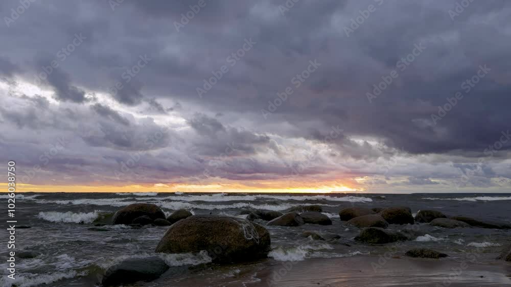 A stunning sunset view of the sea, with waves crashing on large boulders along a sandy beach. Shot from a low angle with the horizon at eye level, capturing the dramatic contrast of the vibrant sunset
