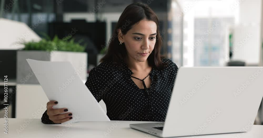 Lebanese businesswoman, office employee sits at desk using laptop, holding papers, review information, preparing agreement documents for negotiations or presentations, researching market looks focused Lebanese businesswoman, office employee sits at desk using laptop, holding papers, review information, preparing agreement documents for negotiations or presentations, researching market looks focused