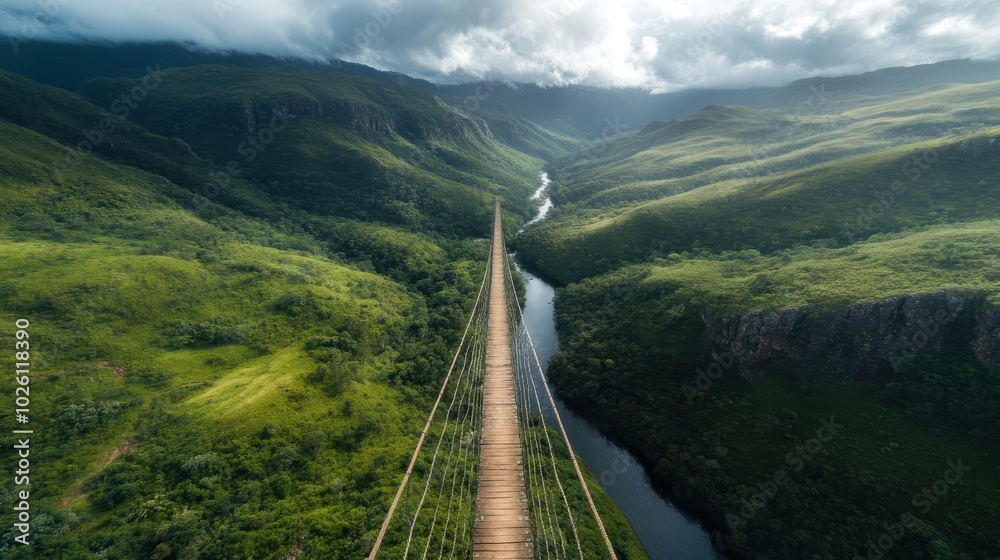 Fototapeta premium A stunning aerial view of a suspension bridge crossing a vibrant green valley, with a river meandering below and mountains receding into the cloudy distance.