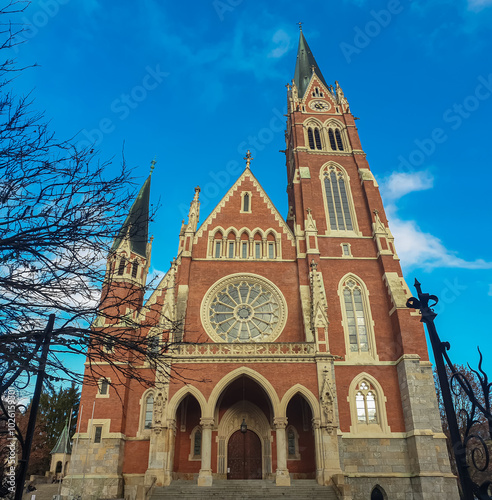 Majestic church Herz Jesu Kirche in Graz, Styria, Austria. Landmark against clear blue sky. Intricate architectural details, including pointed spire, ornate windows, clock face. Lush green foliage photo