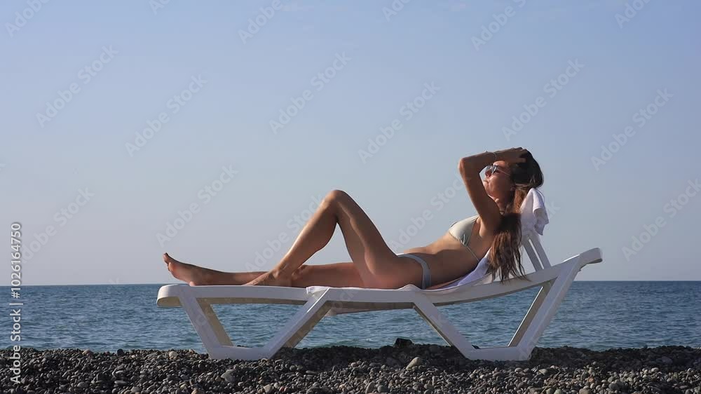A young woman relaxes on a beach lounge chair by the ocean, sipping a refreshing drink. She enjoys the sunny weather and peaceful atmosphere, perfect for a summer vacation by the sea. A young woman relaxes on a beach lounge chair by the ocean, sipping a refreshing drink. She enjoys the sunny weather and peaceful atmosphere, perfect for a summer vacation by the sea.