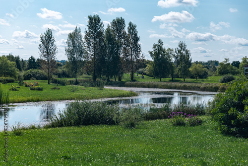 Pond reflecting a bright blue sky with fluffy clouds. The pond is surrounded by lush green grass and trees, creating a peaceful and serene atmosphere. The image evokes a sense of serenity. photo