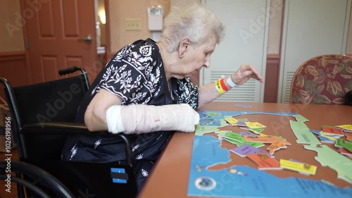 An elderly woman with a broken arm, wearing a fall risk bracelet, sits in a wheelchair and assembles a colorful US state puzzle on a table. photo