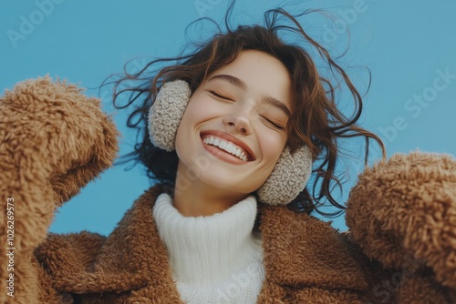 Young woman happily enjoying music while wearing cozy earmuffs against a blue...