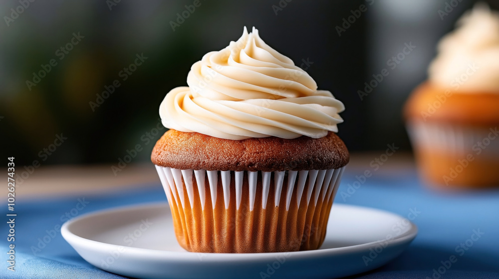 Close-up of a vanilla cupcake with smooth cream icing on a white plate, with another cupcake in the background, all placed on a blue tablecloth. Close-up of a vanilla cupcake with smooth cream icing on a white plate, with another cupcake in the background, all placed on a blue tablecloth.