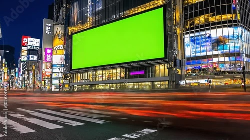 A city street at night with a green screen billboard, illuminated by streetlights. The street is blurred, showing motion of cars.