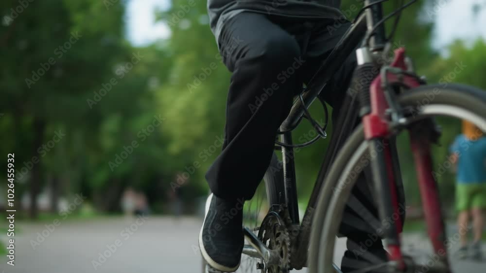 Young boy riding a bicycle steadily through a park area, wearing glasses and a grey top, the background includes lush view of trees, an urban building, and blurred people walking in the background Young boy riding a bicycle steadily through a park area, wearing glasses and a grey top, the background includes lush view of trees, an urban building, and blurred people walking in the background