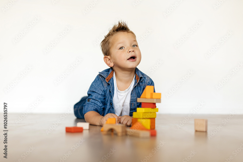 A cheerful boy lays on his stomach, engaging with vibrant wooden blocks as he constructs a tower. Natural light fills the room, enhancing the playful atmosphere. A cheerful boy lays on his stomach, engaging with vibrant wooden blocks as he constructs a tower. Natural light fills the room, enhancing the playful atmosphere.