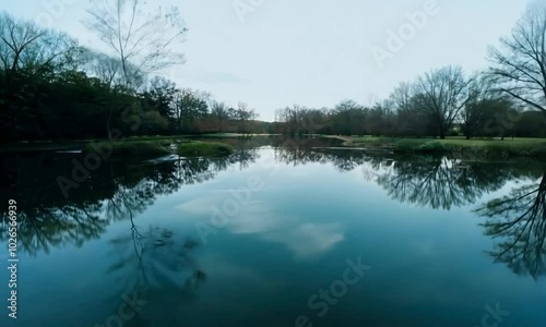 Tracking shot with reflected scenery: A clear, reflective pond with surrounding trees and sky mirrored in the water. The camera tracks along the pond’s edge, capturing the reflections and the transpar photo