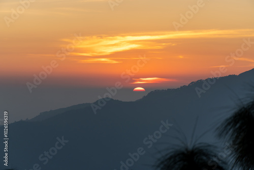 Sun is sinking behind the horizon, Painting sky in warm orange and gold. Mountain ridge covered in dark forest silhouette and blurred pine treetops in the foreground beneath the dark orange clouds. 