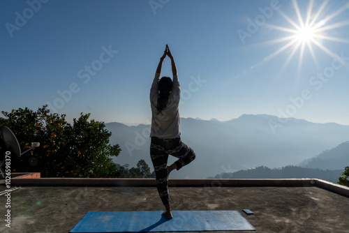 A young woman holds a yoga pose on yoga mat.Sunrise silhouette against layered dusky mountains.The morning light casts sun flares, adding a serene glow to her peaceful practice.Lifestyle in mountains.