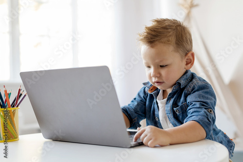 Online Education. A young boy is intently working on a laptop at a bright indoor table, surrounded by colorful pencils in a container, showcasing a moment of concentration.