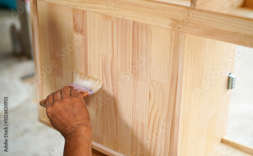 Carpenter hands applying varnish to a cabinet with a paintbrush. First lacquer coating on wood surface at a furniture and wooden workshop.Close up.