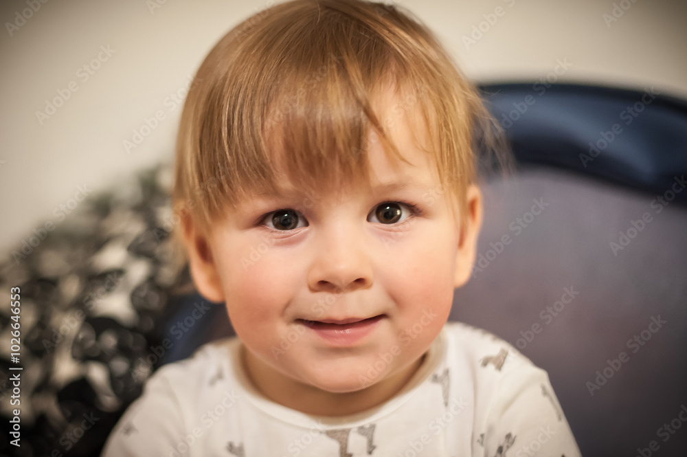 Young child with light brown hair sits smiling in cozy indoor setting wearing a playful patterned outfit in warm lighting during afternoon Young child with light brown hair sits smiling in cozy indoor setting wearing a playful patterned outfit in warm lighting during afternoon