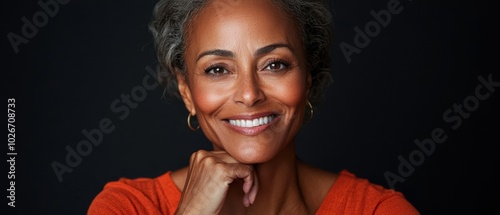  Aperson, smiling directly at the camera, with one hand resting under their chin against a black backdrop photo