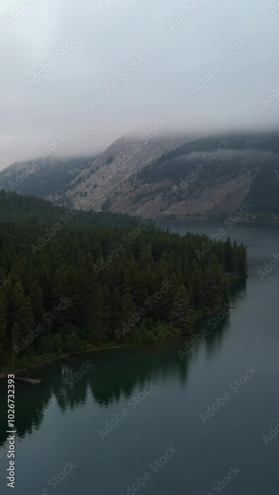Aerial view of a pristine lake reflecting a dense forest and a misty mountain, showcasing the serene beauty of nature. Crowsnest Pass, Alberta, Canada. Aerial view of a pristine lake reflecting a dense forest and a misty mountain, showcasing the serene beauty of nature. Crowsnest Pass, Alberta, Canada.