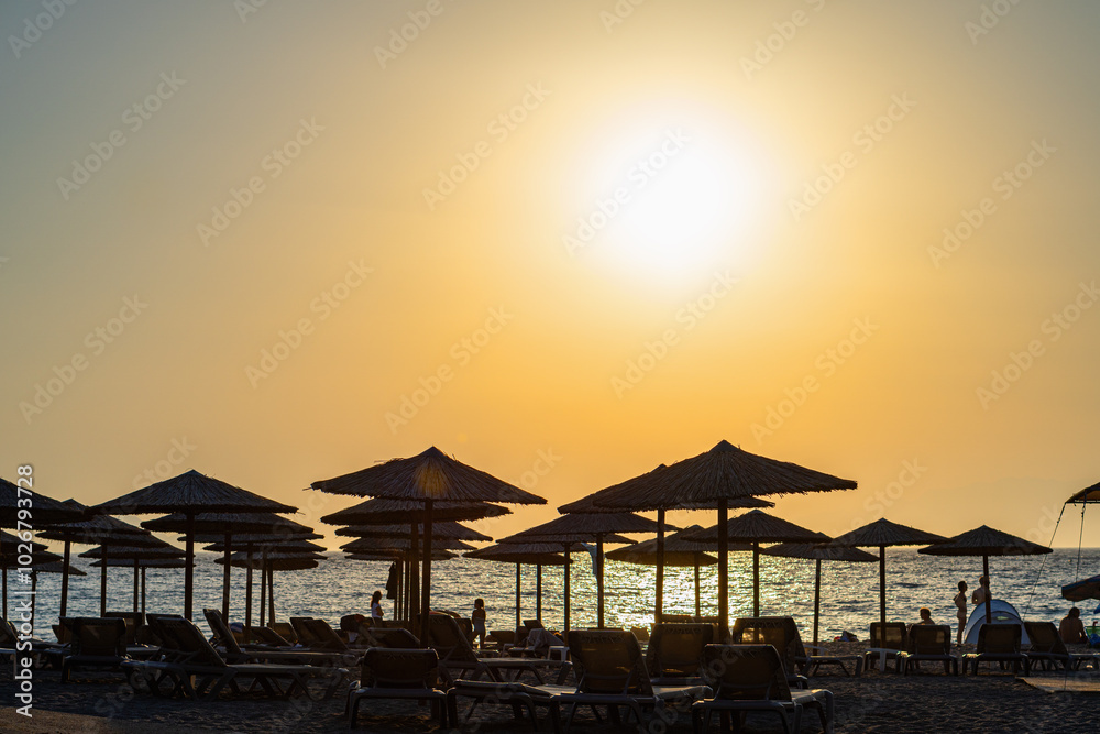Silhouette of umbrellas and sunbeds on seaside beach with yellow sky, sun shining in front, backlit, telephoto Silhouette of umbrellas and sunbeds on seaside beach with yellow sky, sun shining in front, backlit, telephoto