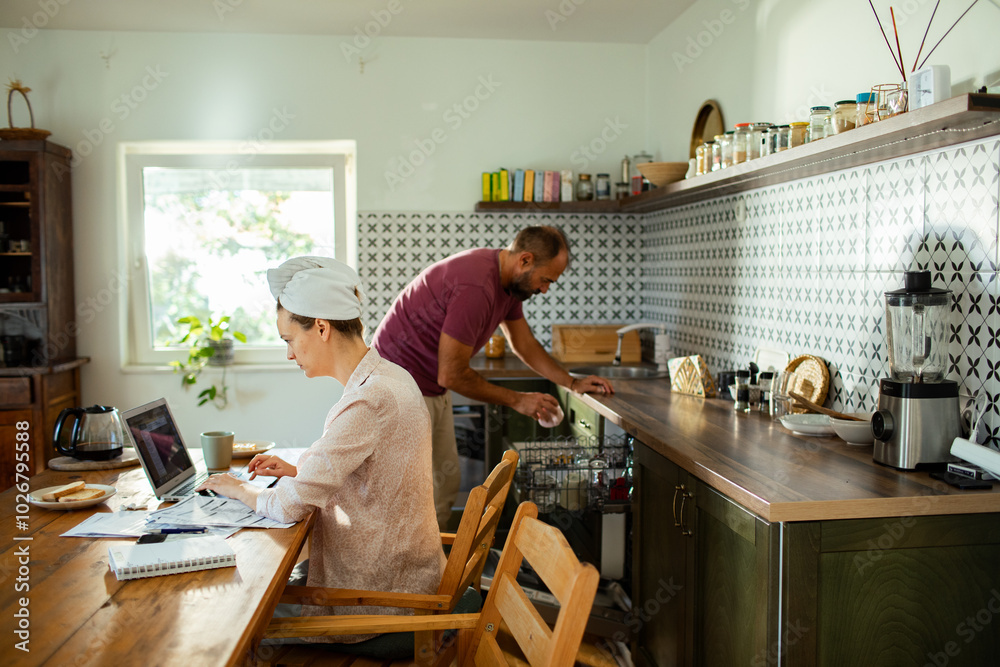 Woman working on laptop with bills while man loads dishwasher in kitchen Woman working on laptop with bills while man loads dishwasher in kitchen