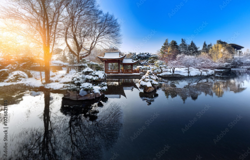 Obraz premium A wooden gazebo sits on the edge of a calm pond, surrounded by snow-covered trees and a clear blue sky.
