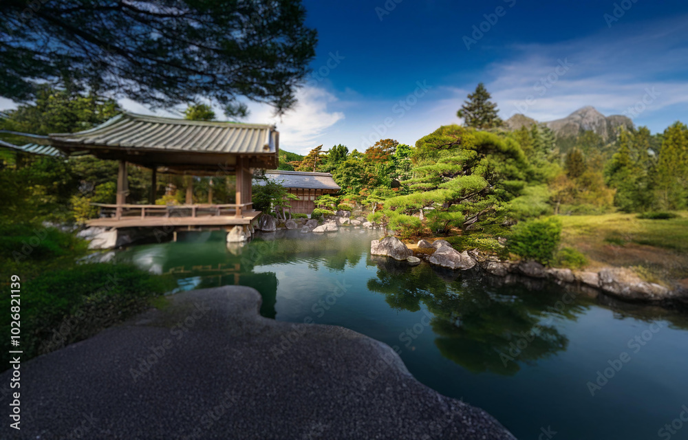 A traditional Japanese pavilion sits on a wooden platform over a still pond surrounded by lush foliage, with a mountain in the distance. A traditional Japanese pavilion sits on a wooden platform over a still pond surrounded by lush foliage, with a mountain in the distance.