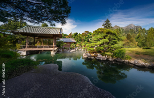 A traditional Japanese pavilion sits on a wooden platform over a still pond surrounded by lush foliage, with a mountain in the distance.