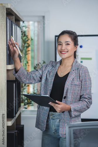 Asian businesswoman is smiling while choosing files in the office shelf and holding a clipboard photo