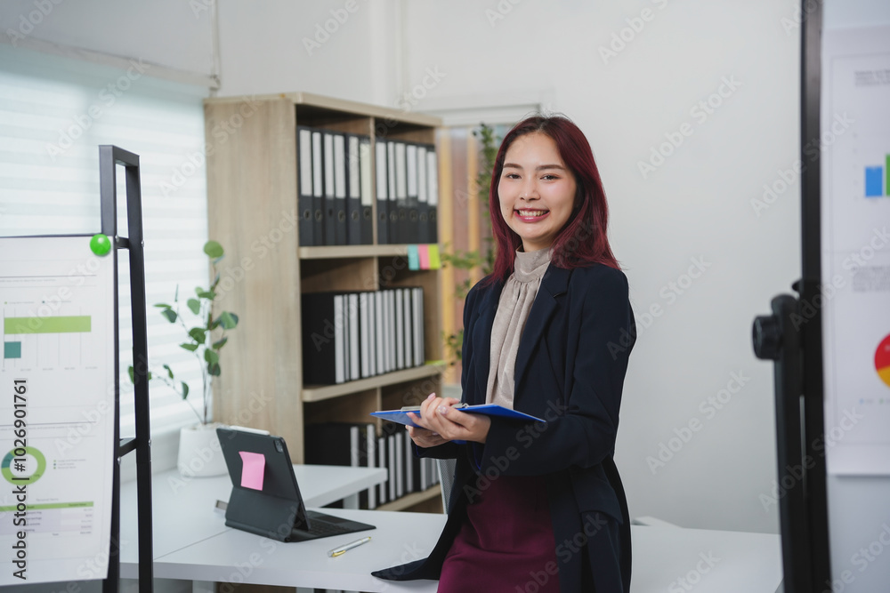 Young manager smiling and holding documents while standing in a modern office with graphs and charts on the wall Young manager smiling and holding documents while standing in a modern office with graphs and charts on the wall
