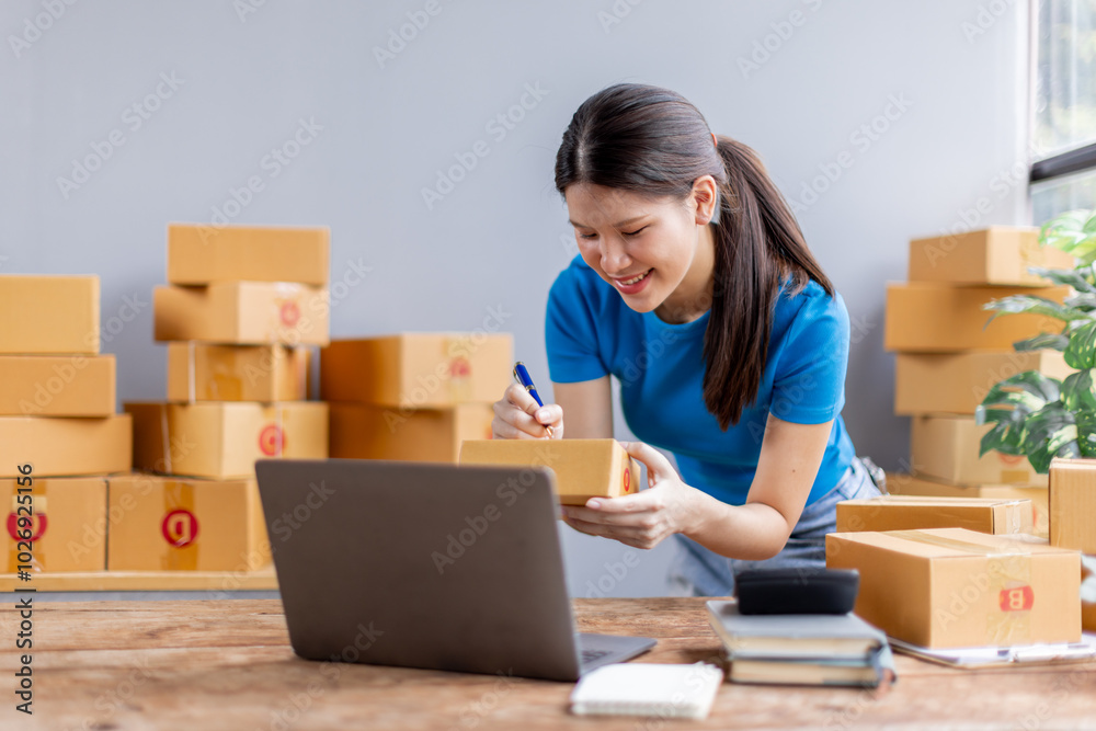 Asian young female sme business owner with product package box at warehouse, merchant seller checking customer address order confirming parcel delivery.
