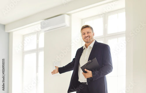 Portrait of happy smiling man realtor agent showing new house standing in white impartments with documents in hands and looking cheerful at camera. Moving day, relocating and mortgage concept