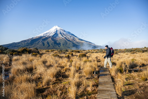 Obraz na płótnie Backpacker hiking the Pouakai circuit on boardwalk among golden tussocks. Mt Taranaki in the background. New Zealand. w salonie