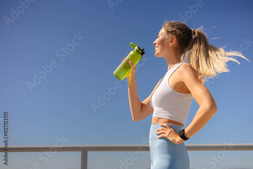 Smiling young blonde woman jogger runner drink water from bottle at river embankment closeup