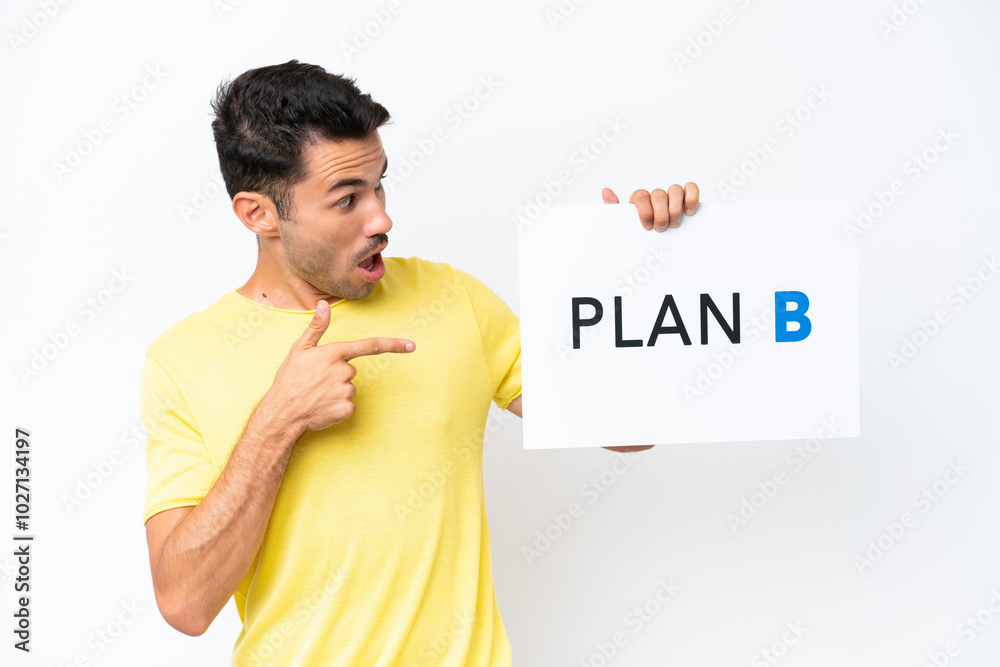 Young handsome man over isolated white background holding a placard with the message PLAN B with surprised expression