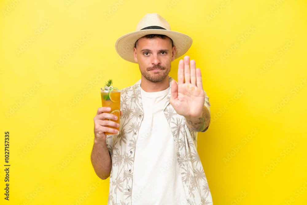 Young caucasian man holding a cocktail isolated on yellow background making stop gesture Young caucasian man holding a cocktail isolated on yellow background making stop gesture