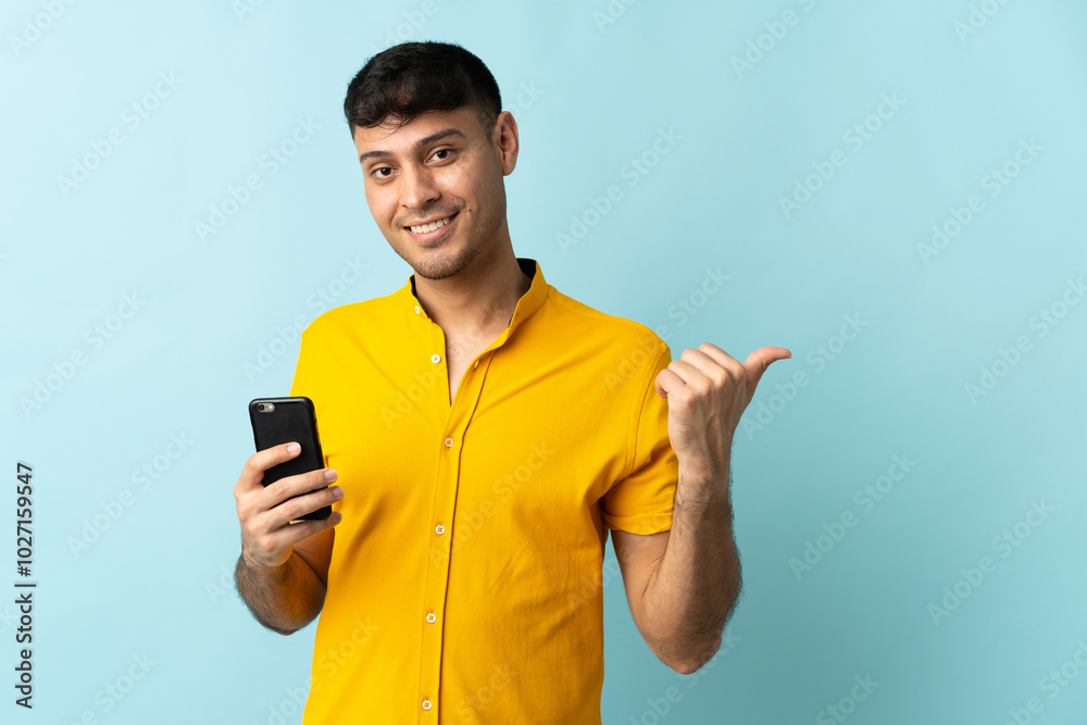Young Colombian man using mobile phone isolated om blue background pointing to the side to present a product Young Colombian man using mobile phone isolated om blue background pointing to the side to present a product