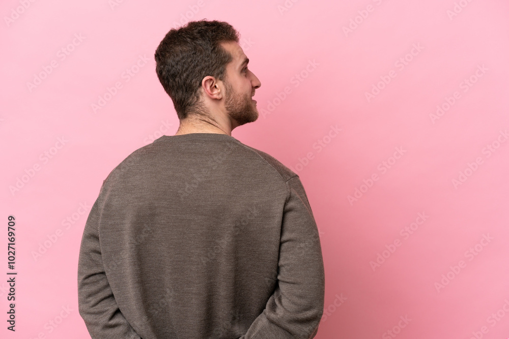 Young caucasian man isolated on pink background in back position and looking back