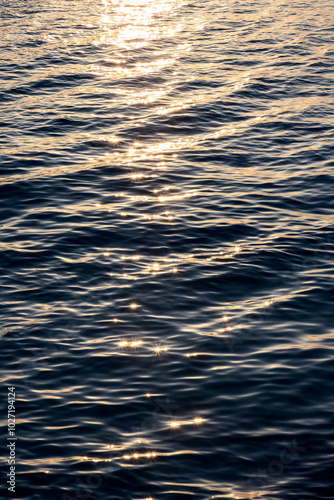 Vertical photo of a textured sea surface at sunset, with ripples reflecting sparkling sun rays. The shimmering water creates a beautiful and dynamic background, perfect for creative projects