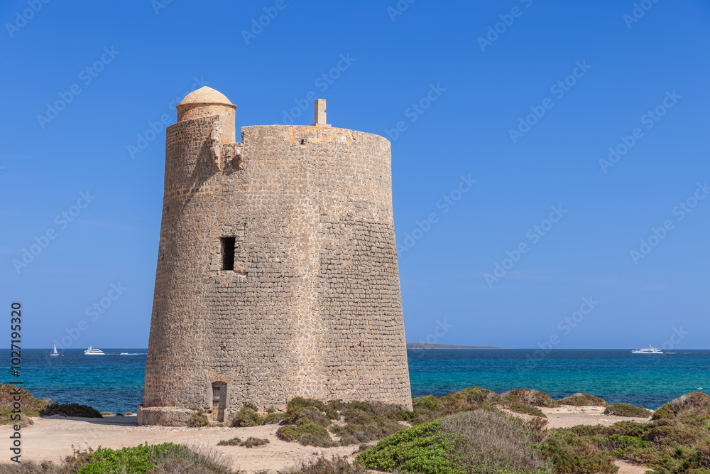 Torre de ses Portes, an ancient watchtower on the Ibiza coast, overlooks the turquoise Mediterranean Sea. The stone structure is set against a vibrant blue sky and framed by coastal plants Torre de ses Portes, an ancient watchtower on the Ibiza coast, overlooks the turquoise Mediterranean Sea. The stone structure is set against a vibrant blue sky and framed by coastal plants