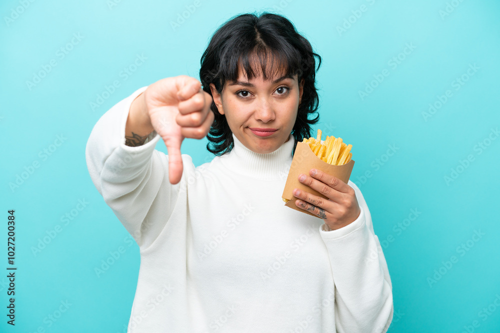 Young Argentinian woman holding fried chips isolated on blue background showing thumb down with negative expression Young Argentinian woman holding fried chips isolated on blue background showing thumb down with negative expression